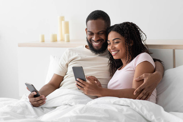 Smiling happy young african american guy hugs lady and typing, surfing, chatting on smartphones on comfortable bed - Foto, afbeelding