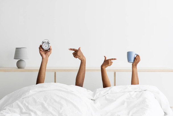 Hands of young black guy and woman stick out from under blankets, holds cups and point finger at alarm clock - Fotó, kép
