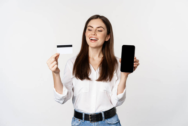 Happy woman showing credit card and smartphone screen, concept of online shopping, buying in app, standing over white background - Foto, imagen