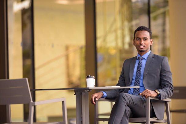 都市でスーツを着てコーヒーショップに座っているハンサムな若いアフリカのビジネスマン - 写真・画像