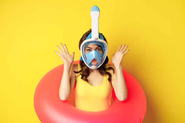Funny happy woman in swimming ring, wearing snorkling mask for diving, posing against yellow background - Фото, изображение