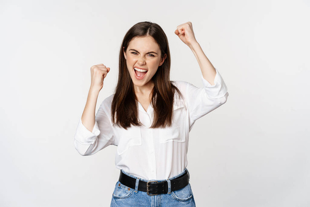 Excited young adult woman reacting to win, surprise news, screaming and cheering, triumphing, achieve goal and celebrating, standing over white background - Φωτογραφία, εικόνα