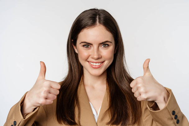Close up portrait of confident businesswoman smiling, showing thumbs up, recommending company, complimenting smth good, standing over white background - Фото, изображение