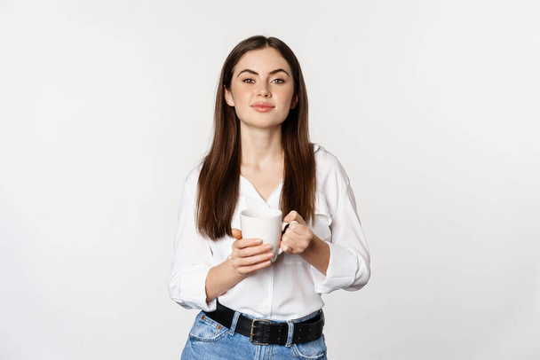 Young moder business woman, office lady holding mug with coffee tea and smiling, standing against white background - Foto, Imagem