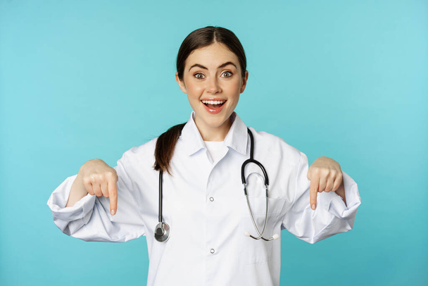 Nadšený zdravotnický pracovník, mladá žena lékař v bílém plášti, ukazuje prsty dolů a usmívá se, ukazuje lékárny reklamy, lékařské promo, modré pozadí - Fotografie, Obrázek