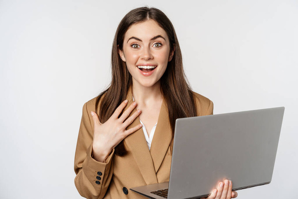 Surprised and excited businesswoman holding laptop, reacting amazed at smth awesome, standing in suit over white background - Photo, Image