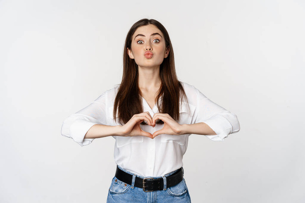 Lovely feminine woman showing heart sign, romantic gesture and smiling with care and tenderness, standing over white background - Fotoğraf, Görsel