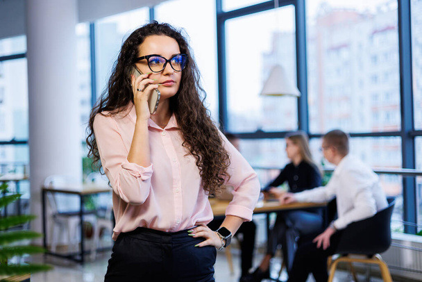 Smart happy young business woman, in stylish clothes and glasses, works in the office, talks with a client on a smartphone, gestures, smiles. Employees are working in the background. Selective focus - Photo, Image