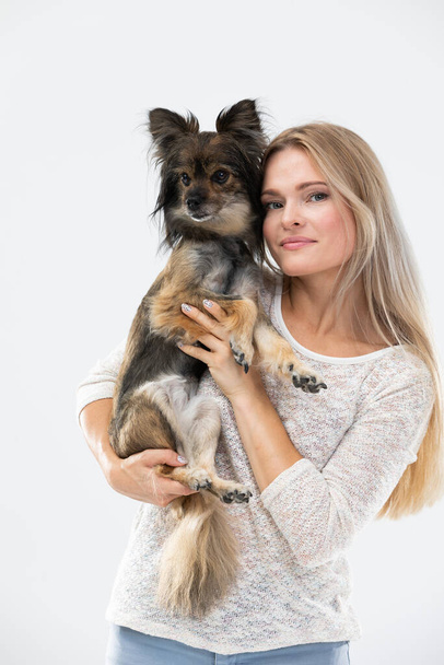 A blonde woman with long hair holds a small dog in her arms and looks at it. Multi-breed dog. - Фото, зображення
