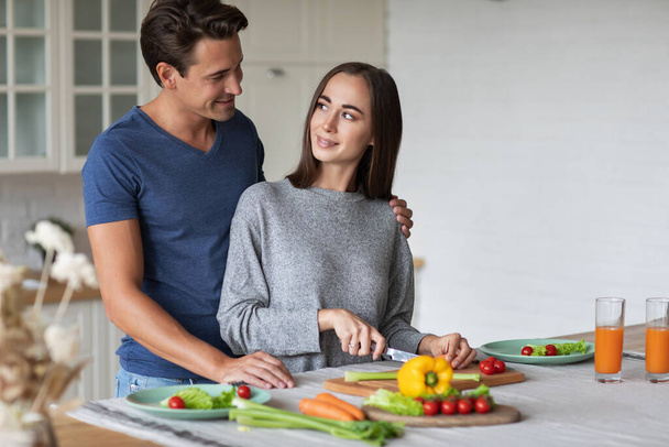 Beautiful young couple cooking vegetable salad while standing in the kitchen at home, enjoying time together. Healthy lifestyle concept. - Фото, зображення