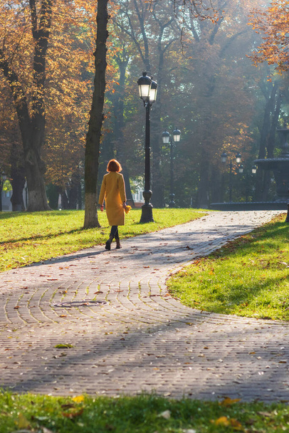 Redhead woman in a yellow coat walks in an autumn park with a bouquet of maple leaves - Фото, изображение