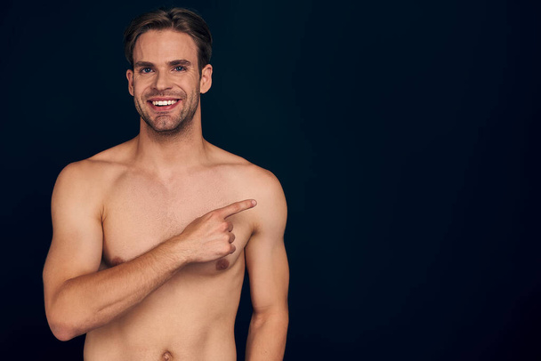 Handsome young man isolated. Portrait of shirtless muscular man is standing on dark blue background and pointing to the side. Showing product - Fotó, kép