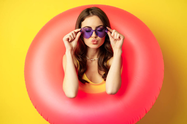 Close up portrait of stylish young woman, girl with swimming ring and summer sunglasses, ready for beach holiday, vacation, standing over yellow background - Foto, afbeelding