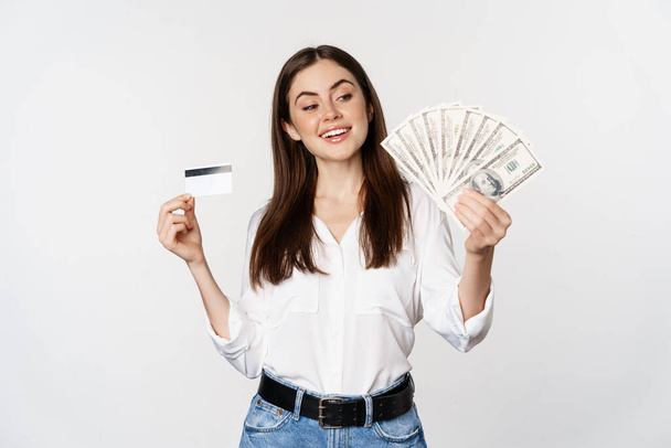 Happy modern woman holding money and credit card in hands, smiling pleased, standing over white background - Fotó, kép