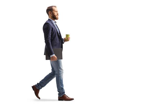 Full length profile shot of a young professional man walking with a takeaway coffee cup and a laptop isolated on white background - Valokuva, kuva