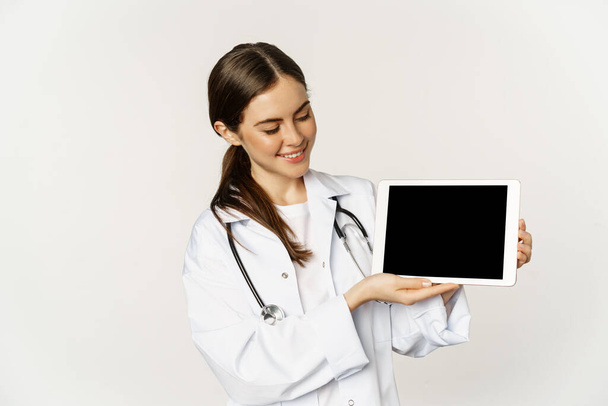 Χαμογελώντας γιατρός, γυναίκα γιατρός ή νοσοκόμα που δείχνει ψηφιακή οθόνη tablet, επίδειξη σε απευθείας σύνδεση ιστοσελίδα, εφαρμογή, στέκεται πάνω από το λευκό φόντο - Φωτογραφία, εικόνα