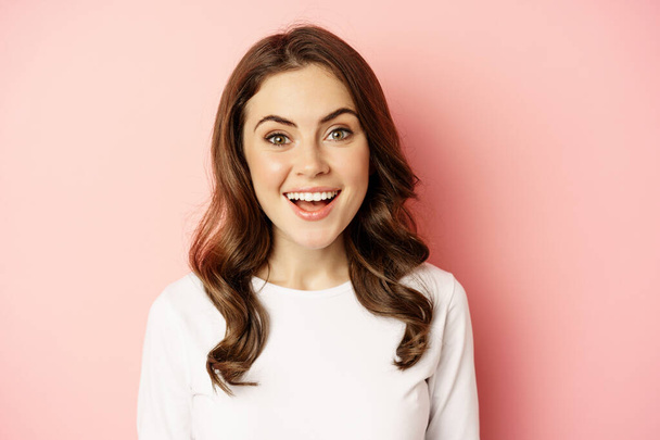 Close up of enthusiastic brunette girl with makeup, smiling and looking happy at camera, posing against pink background - Zdjęcie, obraz