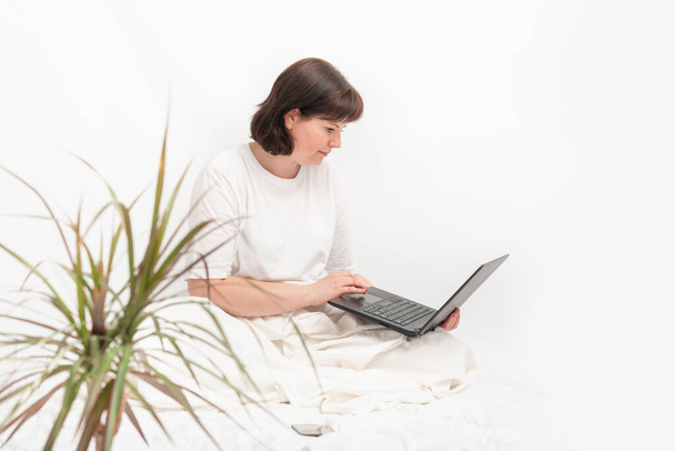Business woman with laptop and phone in bed against a white wall. Young woman works sitting in bed. - Photo, Image