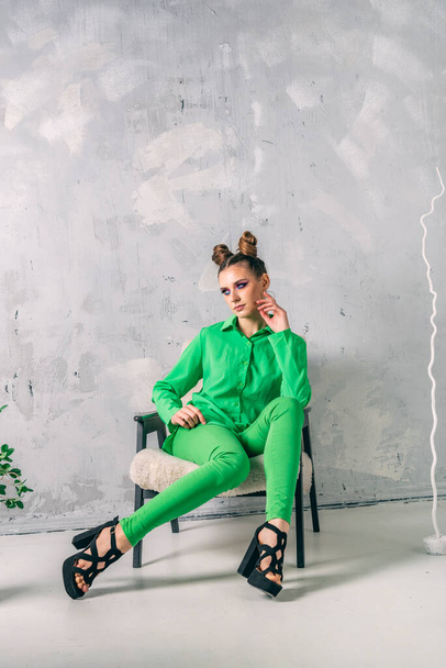 現代のスタジオでプロのファッションモデルを撮影。緑のスーツのスタイリッシュな女性は灰色の背景に椅子に座っています。縦枠. - 写真・画像
