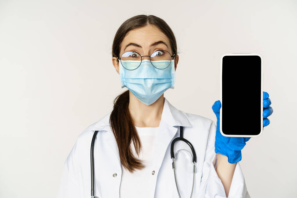 Концепция медицинской помощи онлайн. Удивлённая женщина-врач в маске для лица, удивлённая, показывая мобильное приложение, сайт смартфона, белый фон - Фото, изображение