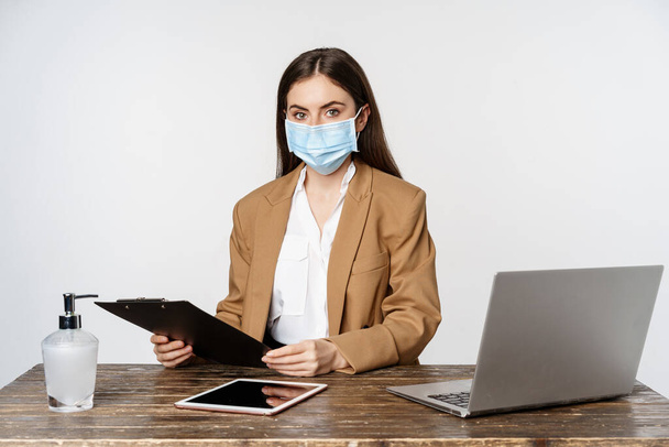 Covid, Pandemie und Büroarbeitsplatzkonzept. Porträt einer Frau mit medizinischer Gesichtsmaske aus Coronavirus, arbeitend, vor weißem Hintergrund stehend - Foto, Bild