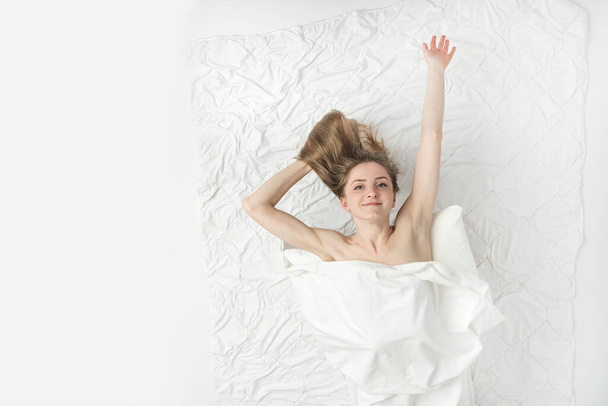 Beautiful naked woman lies in bed hands up, covered with white sheet and enjoys the weekend. Relaxing at home. White background. Top view. - Foto, imagen