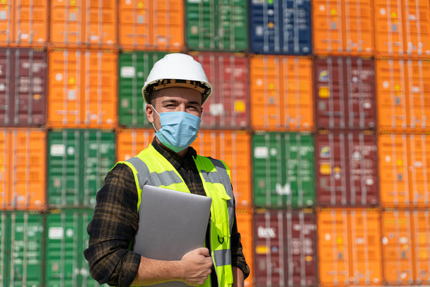 Porträt eines Logistikingenieurs mit medizinischer Gesichtsmaske, der während der Pandemie draußen auf einem großen Handelsdock steht - Foto, Bild