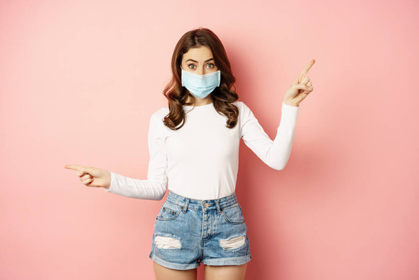 Portrait of beautiful woman in face medical mask pointing sideways, showing two choices, ways, sides, demonstrating advertisement, pink background - Фото, изображение