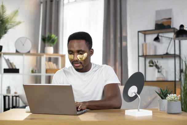 A young man performs cosmetic procedures using gold patches while typing on a laptop - Fotó, kép