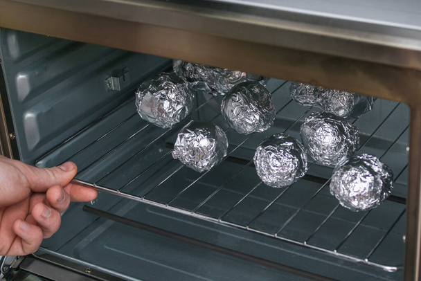 A man puts raw potatoes wrapped in foil on wire rack in the oven. - Фото, зображення