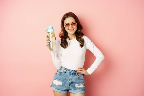 Стильна дівчина в весняному вбранні, одягнена в сонцезахисні окуляри, тримає пляшку води з лимоном, здоровий напій, сміється і посміхається, стоїть над рожевим тлом
 - Фото, зображення