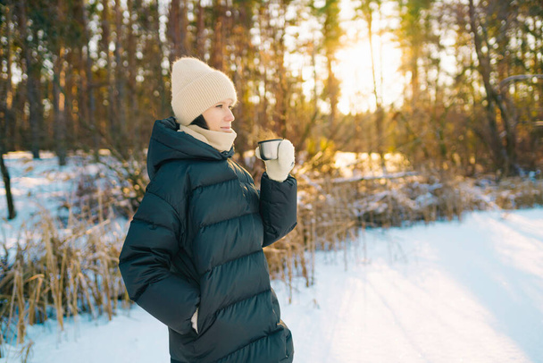 Close up shot of young beautiful woman drinking hot tea from metal mug outdoors on winter day - Photo, image