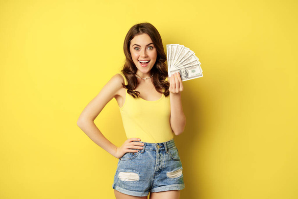 Shopping, credit and money concept. Young brunette woman showing cash and smiling pleased, standing over yellow background - Foto, afbeelding
