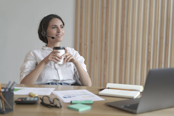 Pleasant italian business woman in headset sits at desk at home office with takeaway coffee - Foto, imagen