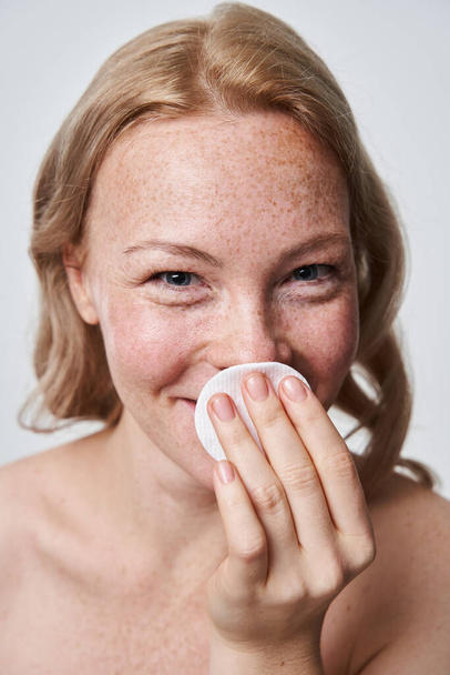 Smiling freckled woman hiding her face behind the cotton pad while removing makeup - Foto, Imagen