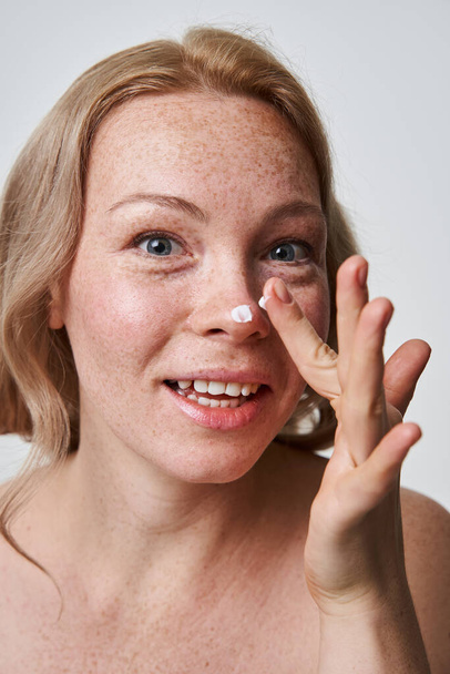 Freckled woman putting moisturizing cream at her nose and smiling - 写真・画像