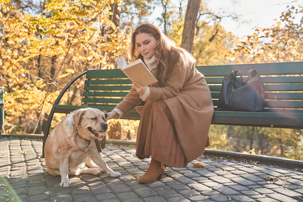 Femme adulte souriante caressant son chien et lisant un livre sur le banc - Photo, image