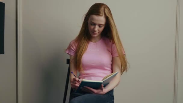 Caucasian woman sitting on the chair at home and writing in her planner book. Red-haired woman. Study day concept. Writing education concept. Lifestyle concept - Záběry, video