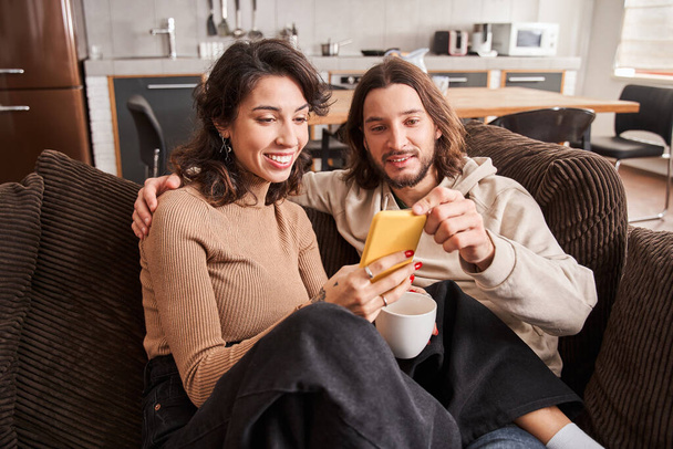 Couple sitting on a couch at home and looking at phone - Photo, Image