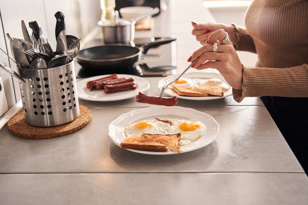 Woman pricks a sausage on a fork over plate with bread and eggs - Foto, Imagem