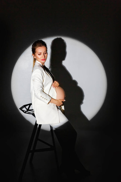 Eine schöne junge Frau im weißen Anzug steht in einem dunklen Studio in einem runden hellen Scheinwerfer.. - Foto, Bild