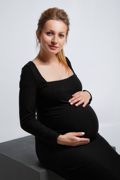Studio portrait for pregnant women of a gorgeous young blonde woman posing on a gray background. A woman sits and gently holds her stomach with her hands - Foto, Bild