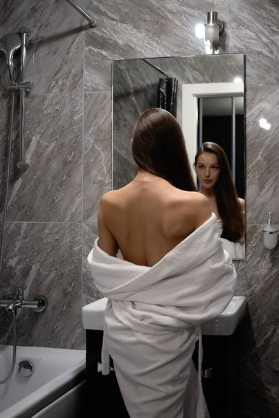 Achteraanzicht van sierlijke sexy vrouw met blote borst staan in de voorkant op spiegel in de badkamer - Foto, afbeelding