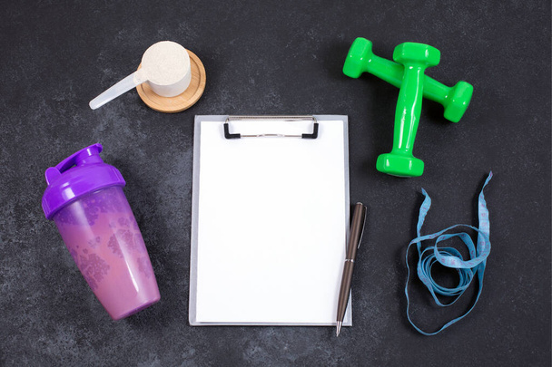 Shaker and scoop of protein powder, dumbbell, measuring tape and notebook on dark table. - Valokuva, kuva