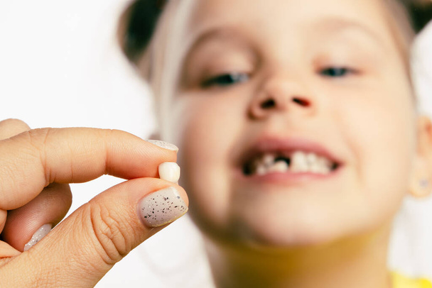 Fingers holding torn out baby milk tooth with little blurred girl face looking at tooth and showing teeth behind on white background. First teeth changing. Going to dentist to do tooth treatment  - Фото, зображення