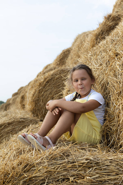 Small cute girl with piece of hay in mouth grimacing and smiling sitting on haystack leaning with back on another stack looking at camera with dozens of haystacks in background. Away from city - Photo, Image