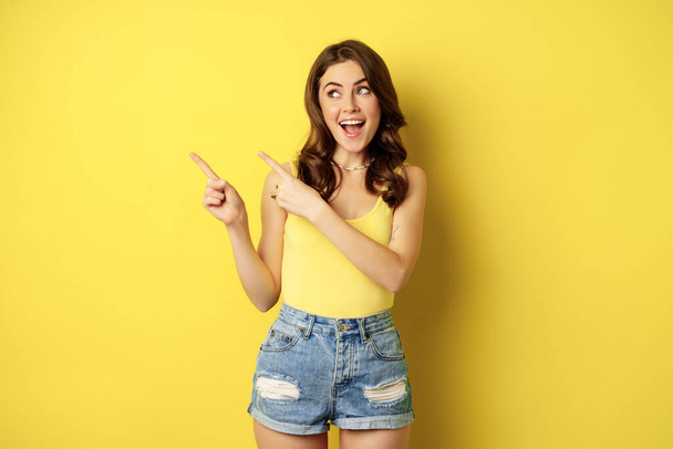 Portrait of stylish summer girl, brunette woman showing way, pointing fingers left, advertising, standing over yellow background - Fotografie, Obrázek