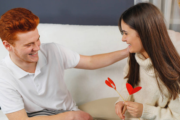 Loving couple sitting on the sofa opposite each other smiling, girl holding red heart cardboard flower and boy put his left hand on her shoulder. - Foto, imagen
