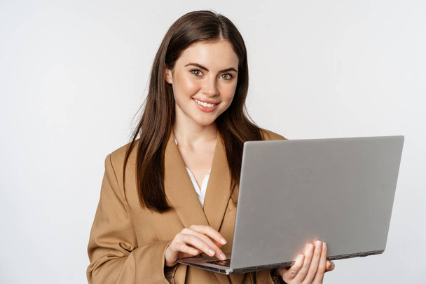 Retrato de la mujer corporativa que trabaja con el ordenador portátil, sonriendo y mirando asertivo, fondo blanco - Foto, imagen