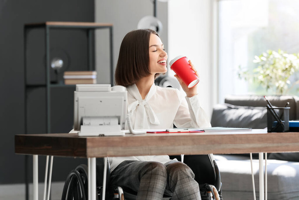Νεαρή γυναίκα με σωματική αναπηρία πίνει καφέ στο γραφείο - Φωτογραφία, εικόνα